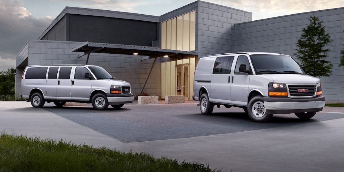Two 2025 GMC Savana vans — a Passenger Van and a Cargo Van — parked outside a modern industrial building.