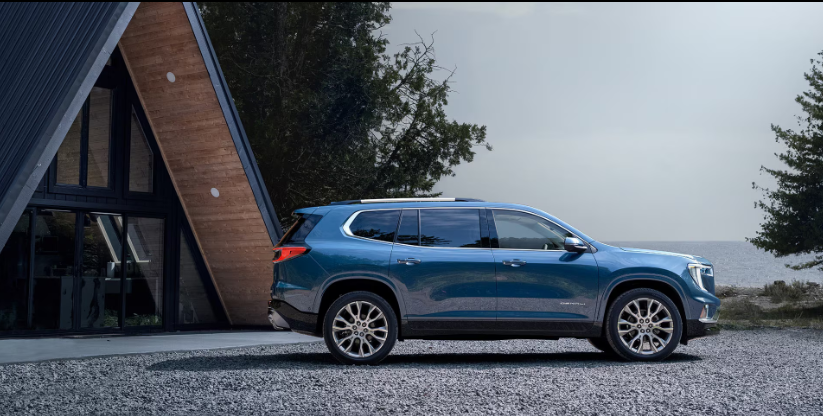 A blue GMC Acadia parked beside a modern A-frame cabin near the water, showcasing its sleek design and upscale styling.